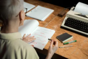Editor reviewing a printed manuscript with red-ink marks at a wooden desk beside a typewriter.