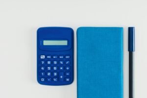 Overhead photo of a blue calculator, spiral notebook, and pen on a white desk.