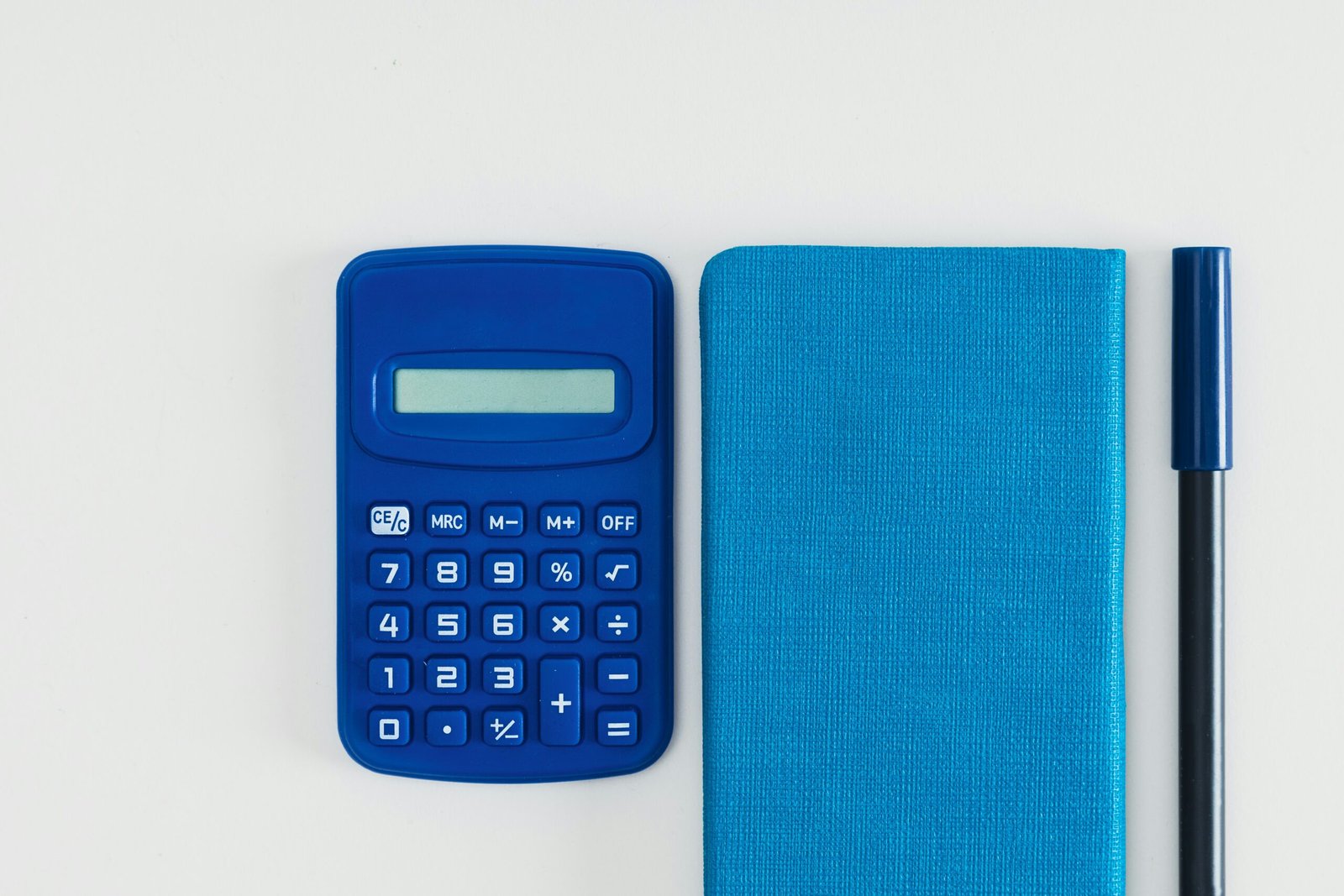 Overhead photo of a blue calculator, spiral notebook, and pen on a white desk.