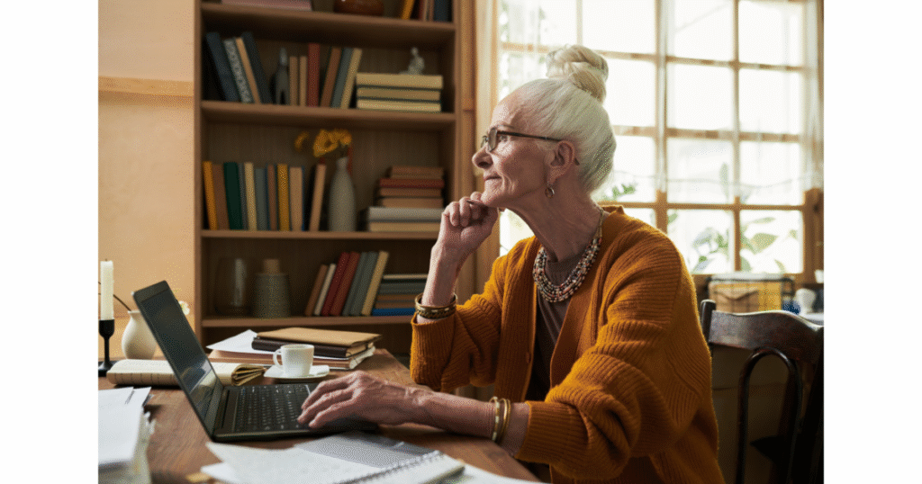 Older woman in a mustard sweater sitting at a desk, reviewing a printed document with a mug beside her.