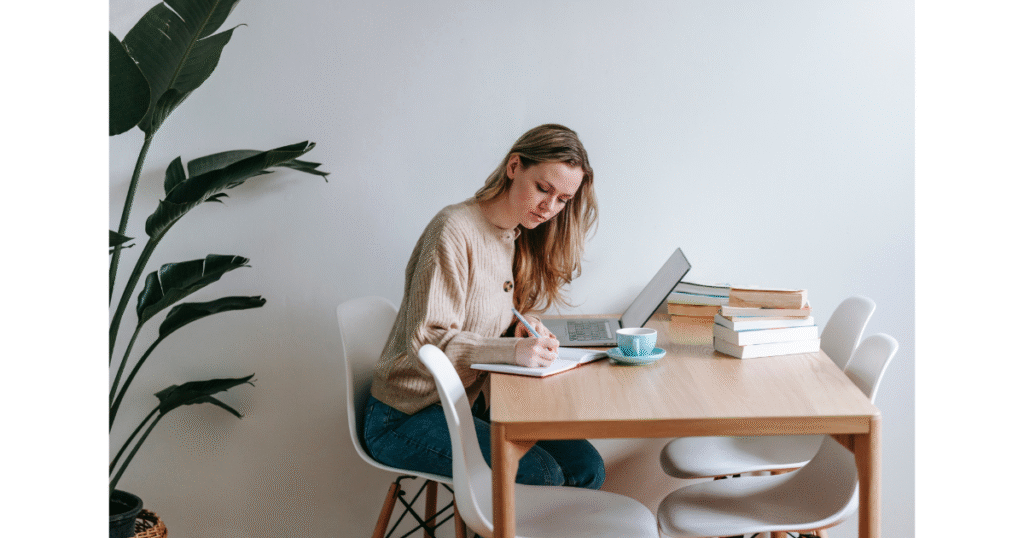 A woman in a light sweater works on a laptop at a white table, surrounded by books and a houseplant