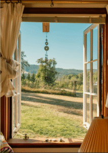 Sunlit meadow seen through an open window with white curtains and a wind chime — a quiet space for debrief reflection.