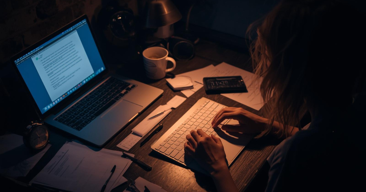 Messy desk with glowing laptop, scattered papers, analog clock showing midnight, and hands typing in a rush.