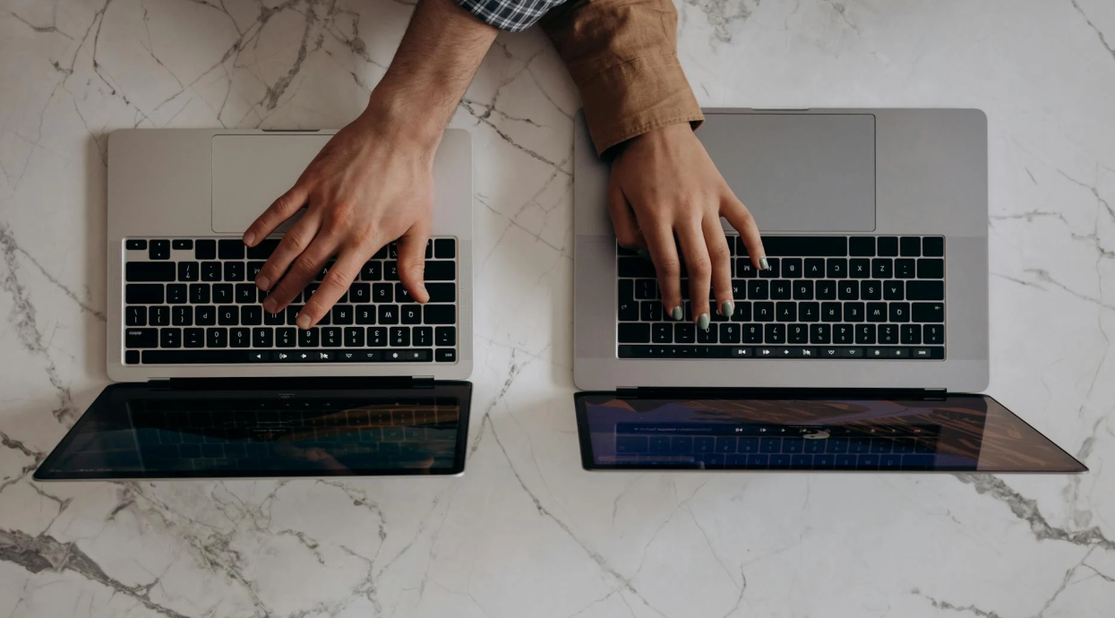 Two people working side by side on laptops at a shared desk, symbolizing collaborative editing and partnership.