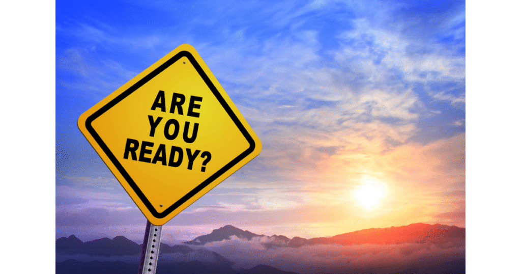 Yellow road sign reading “ARE YOU READY?” in front of a sunrise over mountains and clouds.
